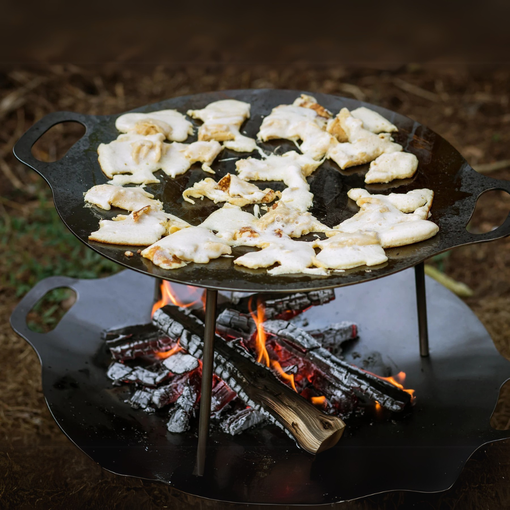 Een ronde Petromax smeedijzeren bakplaat op pootjes boven een kampvuur, gevuld met sissende hamburgers en groenten in de natuur.
