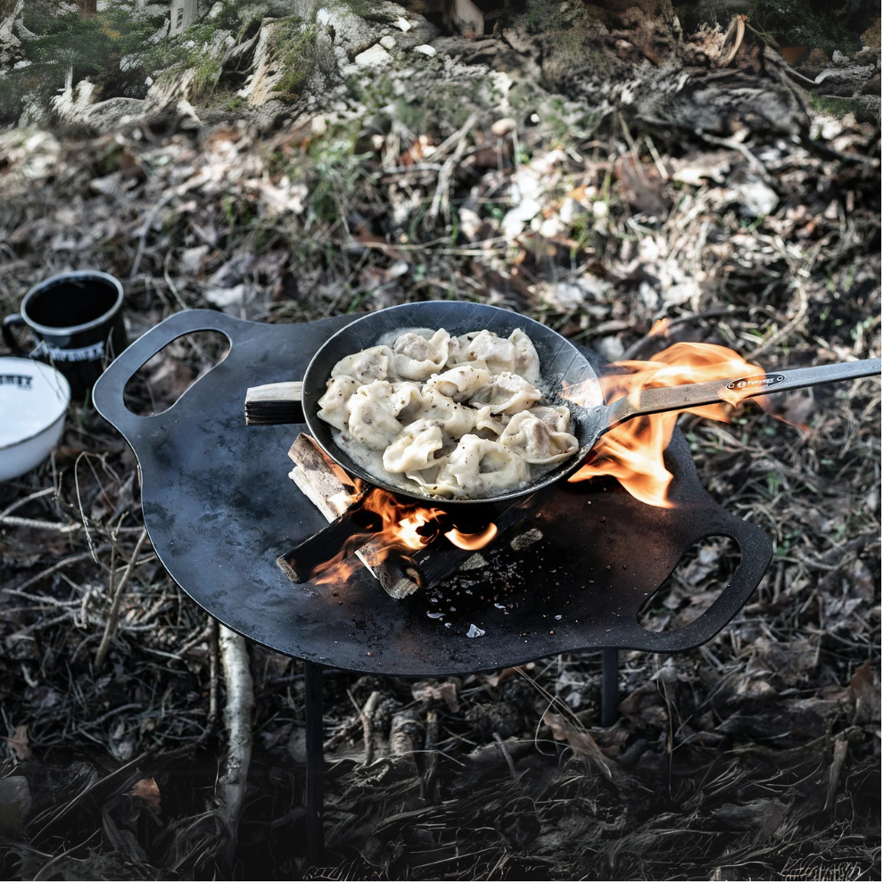 Een ronde Petromax smeedijzeren bakplaat op pootjes boven een kampvuur, gevuld met sissende hamburgers en groenten in de natuur.