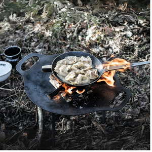 Een ronde Petromax smeedijzeren bakplaat op pootjes boven een kampvuur, gevuld met sissende hamburgers en groenten in de natuur.
