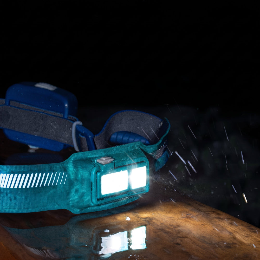 Close-up van de brandende BioLite HeadLamp 440 hoofdlamp in het donker, met de voorlamp die reflecteert op een nat houten oppervlak.