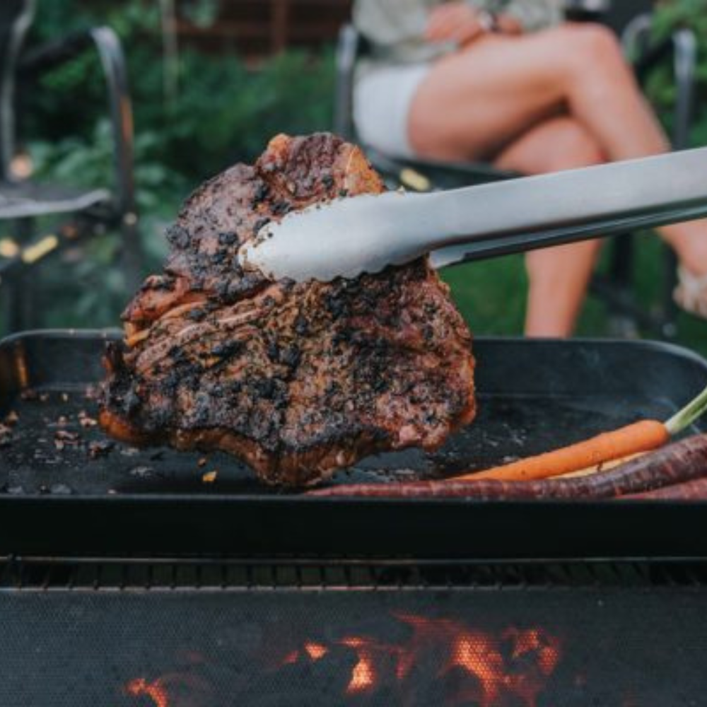 BioLite FirePit+ rvs barbecue grillrooster waarop vlees wordt omgedraaid met een tang boven de vuurkorf