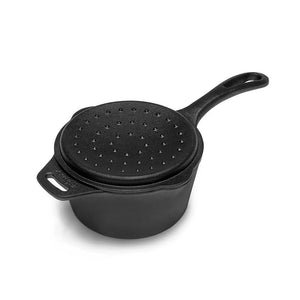 Een zwarte gietijzeren Petromax sauspan met deksel die te midden van gloeiende kolen staat tijdens een outdoor kookbijeenkomst.
