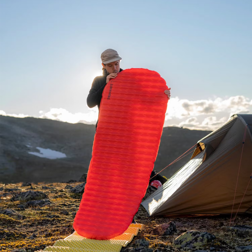 Persoon rolt de rode Therm-a-Rest ProLite Apex Heat Wave L slaapmat uit op de grond bij een tent in de bergen