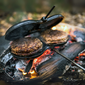 Petromax gietijzeren hamburgerijzer met lange stelen, houten handgrepen en een geribbelde binnenzijde voor het grillen van burgers boven open vuur.