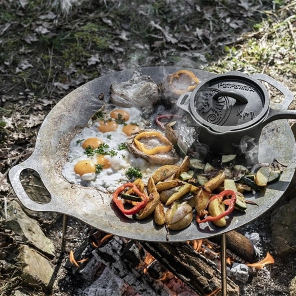 Een zwarte gietijzeren Petromax sauspan met deksel die te midden van gloeiende kolen staat tijdens een outdoor kookbijeenkomst.
