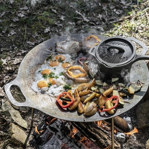 Een zwarte gietijzeren Petromax sauspan met deksel die te midden van gloeiende kolen staat tijdens een outdoor kookbijeenkomst.